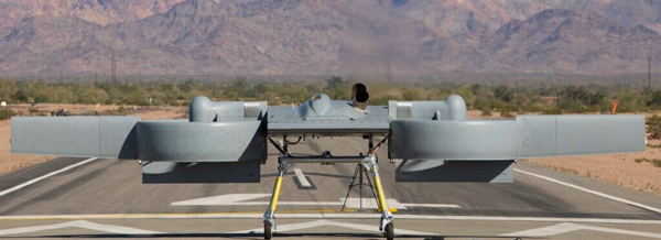 A grey unmanned aerial vehicle sits on a runway with mountains and desert landscape in the background.